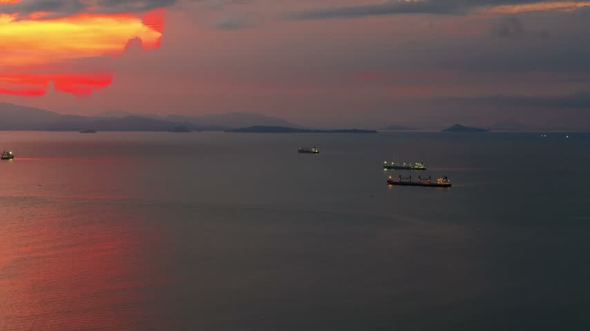 Gorgeous sunset view over the ocean with several large cargo ships sailing on the calm sea while colorful clouds reflect on the water