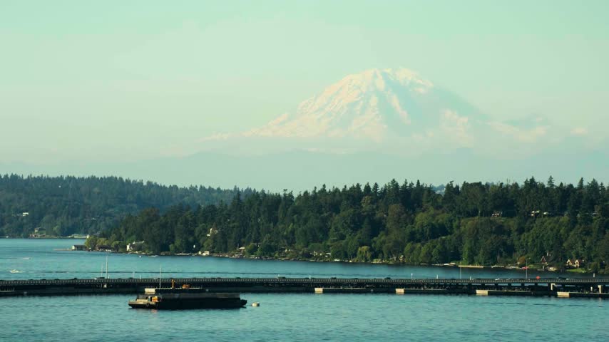 The image shows an aerial view of snow-capped Mount Rainier rising above Lake Washington and a floating bridge. The high-angle drone composition captures the prominent volcanic peak, expansive lake waters, bridge infrastructure, and regional Pacific Northwest geography under daylight conditions. This regional American geographical and transportation asset is ideal for travel documentaries, regional features, and cinematic high-altitude aerial establishing sequences.   