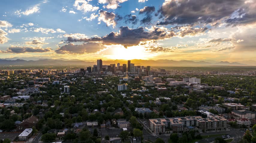 Aerial view of the Denver skyline at sunset with golden light reflecting off buildings, surrounded by urban greenery and distant mountains in Denver, Colorado, United States.