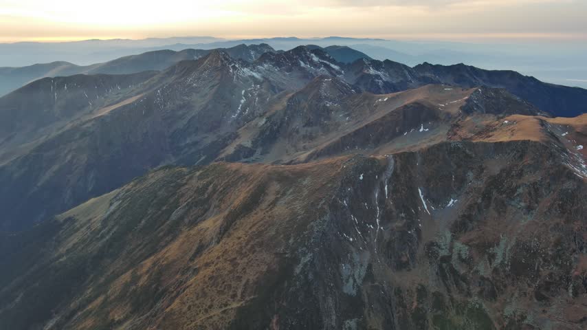 Aerial view of the rugged Transfagarasan mountain range with rocky peaks and patches of snow under soft golden hour light in Cartisoara, Sibiu, Romania.