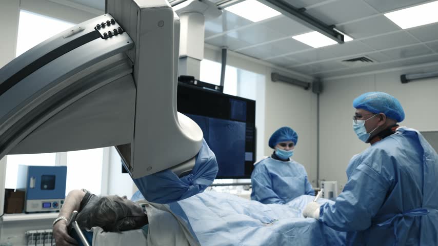 Patient lying under a large C-arm system during an angiography procedure with a medical team.
