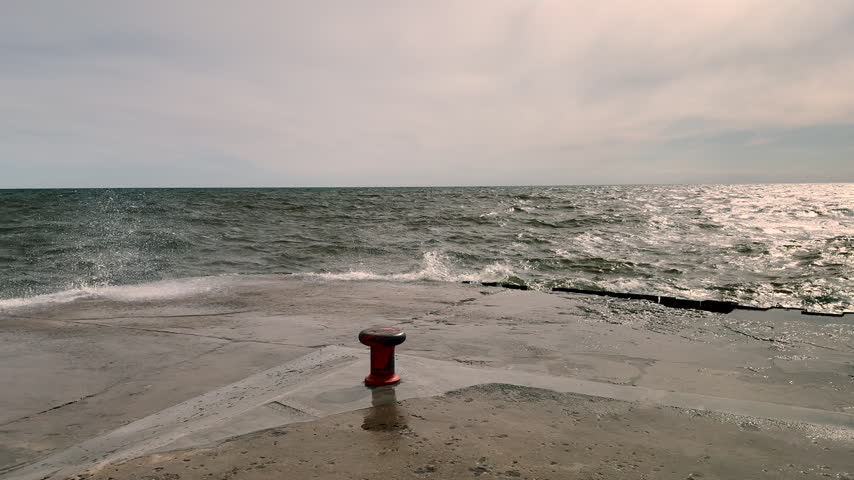 Crashing Lake Michigan Waves Splashing Over Concrete Pier and Bollard. Choppy Great Lakes water and whitecap waves washing over a concrete pier and mooring bollard. Highlights coastal storm energy, nautical docking hardware, and Michigan lakefront scenery.