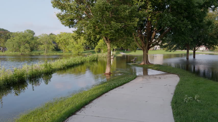 Flooded suburban park and walking path after heavy rainfall, with overflowing lake water covering grass areas.