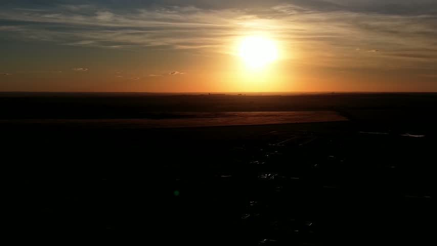 Cinematic aerial view during a dramatic golden sunset, with the drone flying over a dark silhouetted landscape as warm orange light illuminates the horizon and layered clouds.