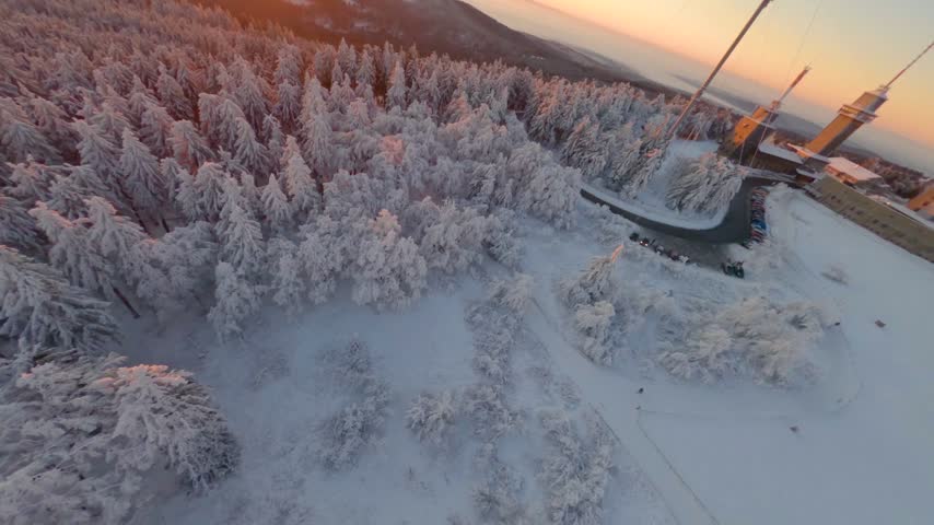 FPV aerial view of a snow-covered forest with frost-laden trees and a communication tower at sunset in Glashutten, Hessen, Germany.