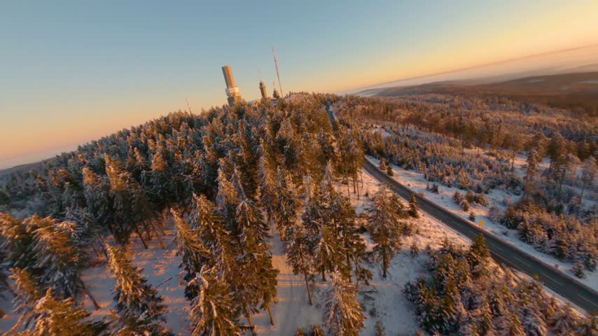 FPV aerial view of a snow-covered forest with a communication tower at sunset in Glashutten, Hessen, Germany.