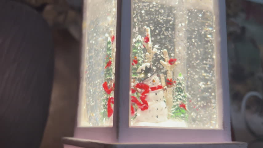 Close-up of a decorative Christmas lantern featuring a cheerful snowman, snowy pine trees and red cardinals surrounded by swirling glitter. Warm glowing light creates a cozy and magical holiday atmosphere, with a dark blurred background and soft bokeh. Festive winter decoration representing Christmas celebration, seasonal warmth, joy and holiday spirit. Ideal for Christmas backgrounds, greeting cards, seasonal advertising, festive campaigns and holiday-themed designs.