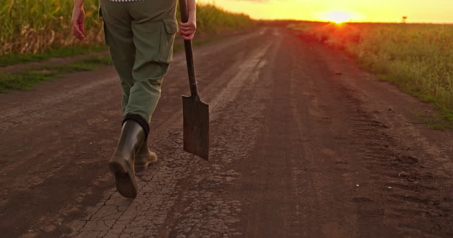 Person in work trousers and rubber boots walks down rural road holding metal spade during warm sunset light. Close up view of footsteps moving along field for farming content.