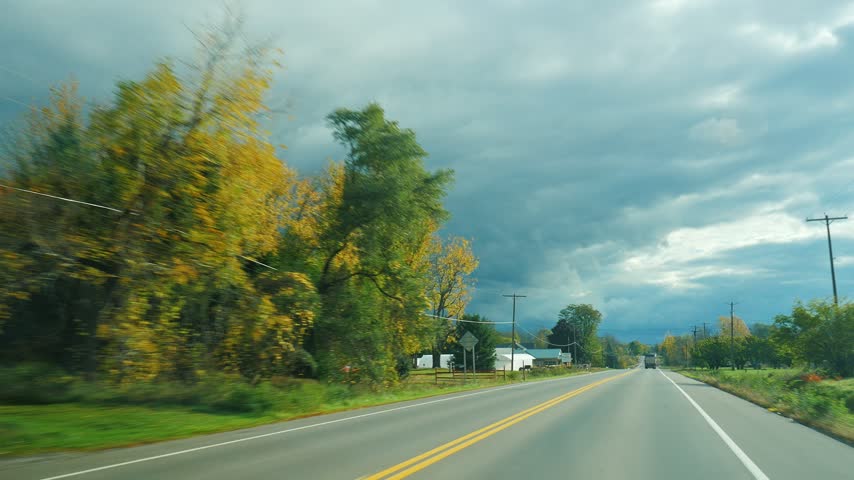 Traveling by car along rural highway past autumn farm market with pumpkins and foliage under dramatic dark storm clouds. Cinematic footage captures seasonal transition scenery.