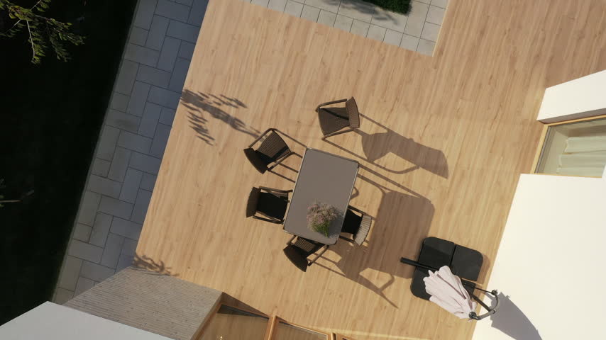Aerial photograph of the courtyard of a modern house. On a spacious wood-look terrace, there is a gray dining table, four wicker chairs, and a closed sun umbrella.