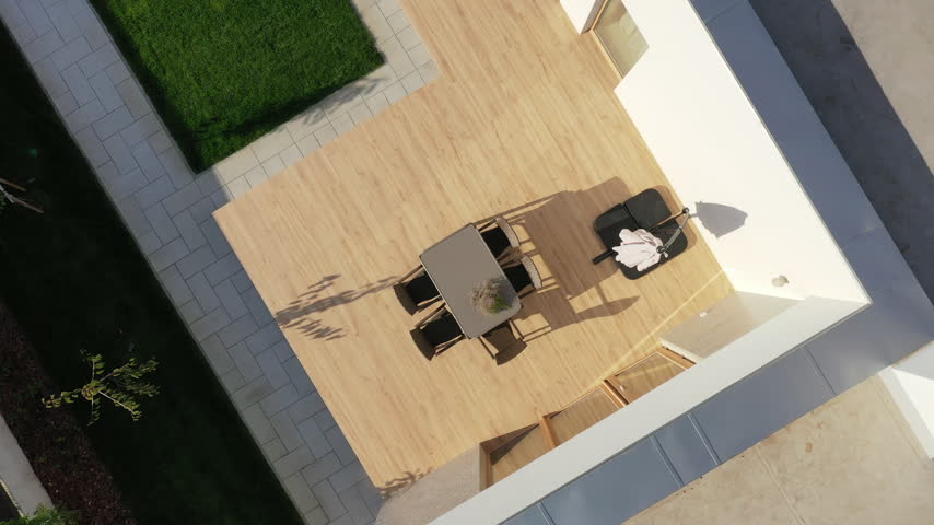 Aerial photograph of the courtyard of a modern house. On a spacious wood-look terrace, there is a gray dining table, four wicker chairs, and a closed sun umbrella.