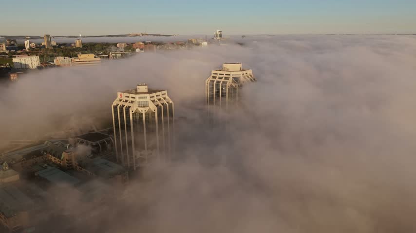 This calm dawn captures Halifax, Nova Scotia, as the rising sun breaks through, illuminating towering buildings amidst a serene fog, creating a picturesque scene.