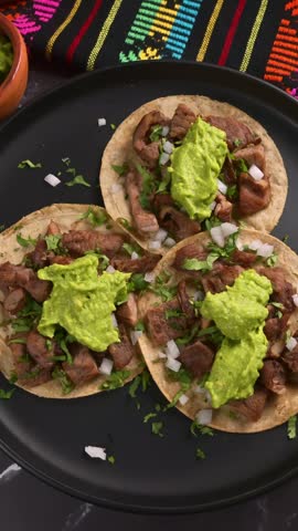 Authentic Mexican beef tacos topped with guacamole, onion and cilantro on a black plate