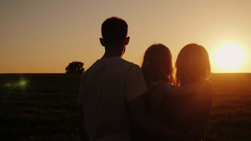 Back view silhouette of father and two daughters standing together in field during warm sunset light. Family bonding outdoors in nature with scenic horizon and golden hour sky.