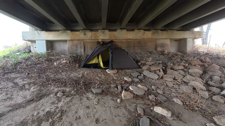 Tent campsite beneath a Historic Route 66 overpass in Sapulpa, Oklahoma, during early spring. A dark outdoor tent with a bright yellow interior is pitched on rocky ground beneath the bridge, surrounded by bridge infrastructure, grassy embankments and early spring vegetation.