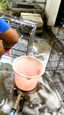 High angle view of a person washing and scrubbing a black wire bird cage with soap foam on a concrete floor.

