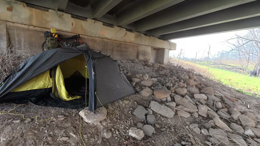 Moving view beneath a Historic Route 66 overpass in Sapulpa, Oklahoma during early spring. The footage shows the underside of the bridge, concrete and steel infrastructure, rocky terrain, grassy embankments and emerging spring vegetation in natural daylight.