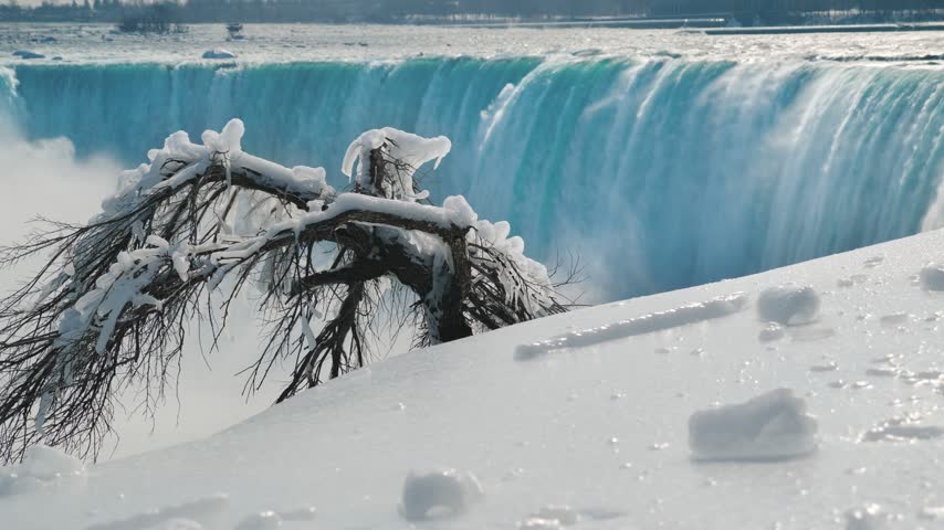 Scenic view of ice covered tree branches in foreground and iconic Niagara Falls with cascading blue water in distance during cold winter season captured in cinematic motion.
