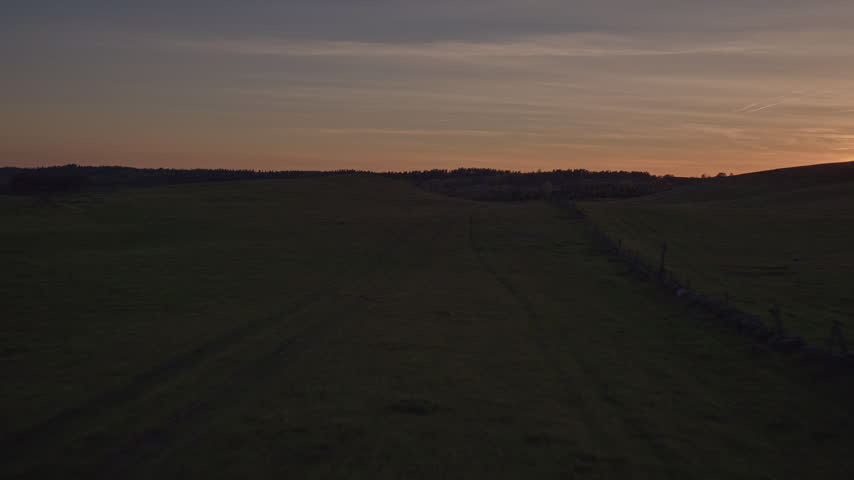 Aerial view of the sun setting behind a forested ridge, casting warm light and lens flare across a silhouetted heath landscape.
