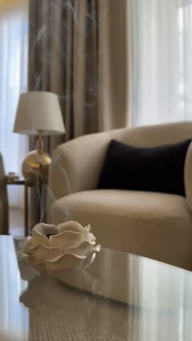 Elegant living room interior featuring a beige armchair with a dark cushion, table lamp, curtains and decorative flower ornament on a reflective glass table.