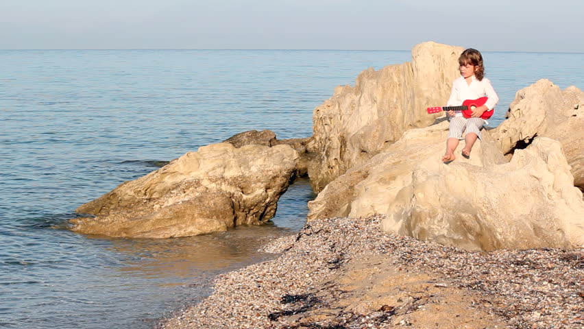 little girl sitting on a rock by the sea and playing guitar