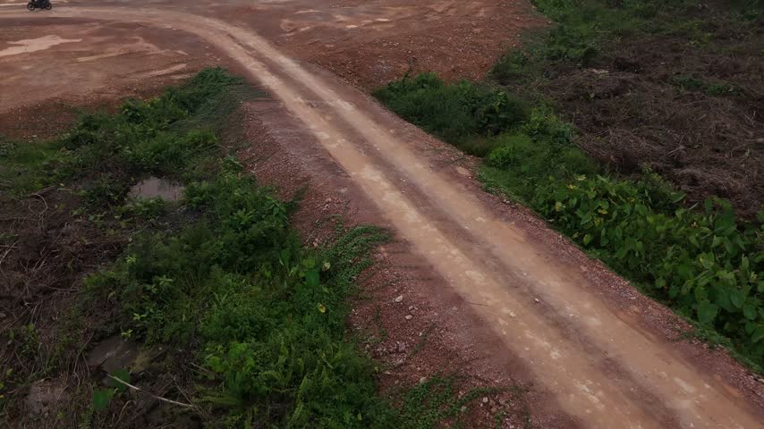 A drone shot showing a red dirt road cutting through a cleared construction site, with gravel, tire tracks, and wild green vegetation.
