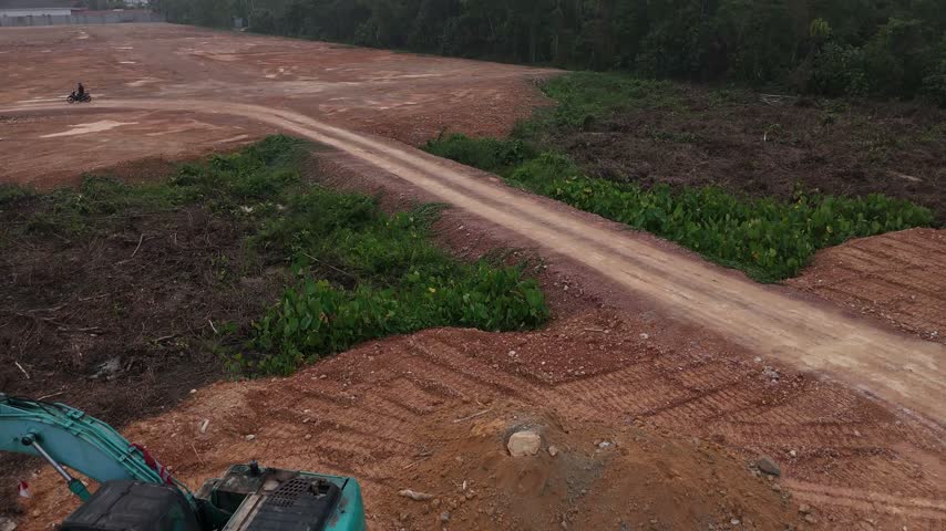 A drone shot showing a red dirt road cutting through a cleared construction site, with gravel, tire tracks, and wild green vegetation.