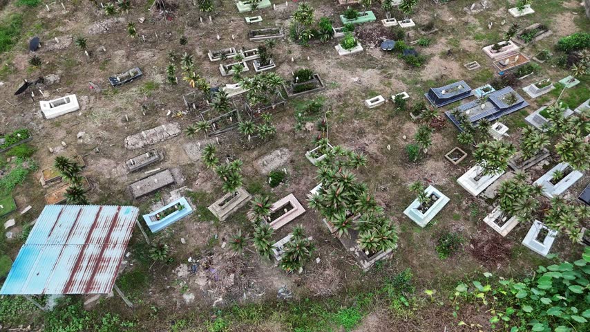 A view from above of a cemetery in Indonesia overgrown with weeds, featuring rectangular graves and a rusty shack.