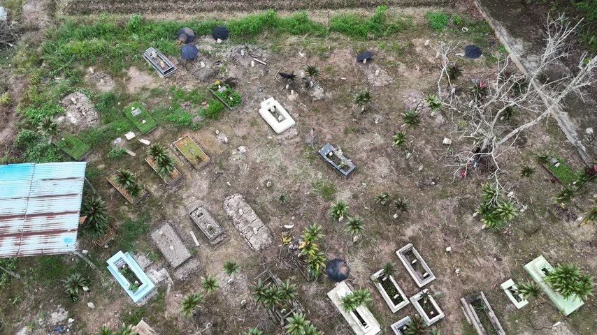 A view from above of a cemetery in Indonesia overgrown with weeds, featuring rectangular graves and a rusty shack.