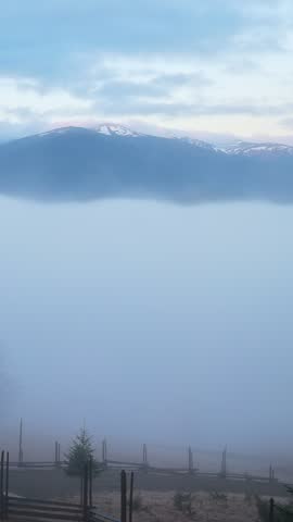 Misty mountain pasture at dawn with quiet forest and distant peaks, soft video frame showing fog drifting over wooden fences, trees and blue ridges