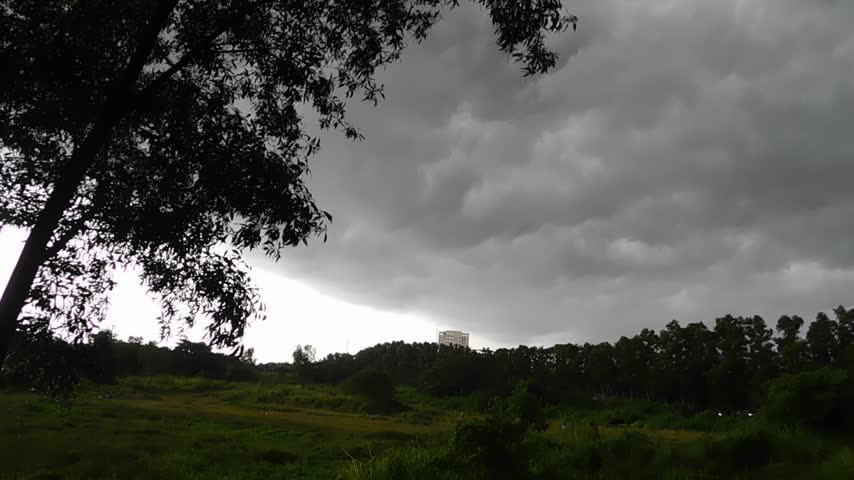 Dramatic rural landscape with dark storm clouds moving across the sky above a green field and tree line, creating a moody natural atmosphere.