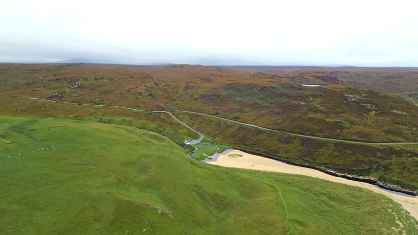 Aerial view rising over grassy hills, sandy beach, and winding rural road in Lewis