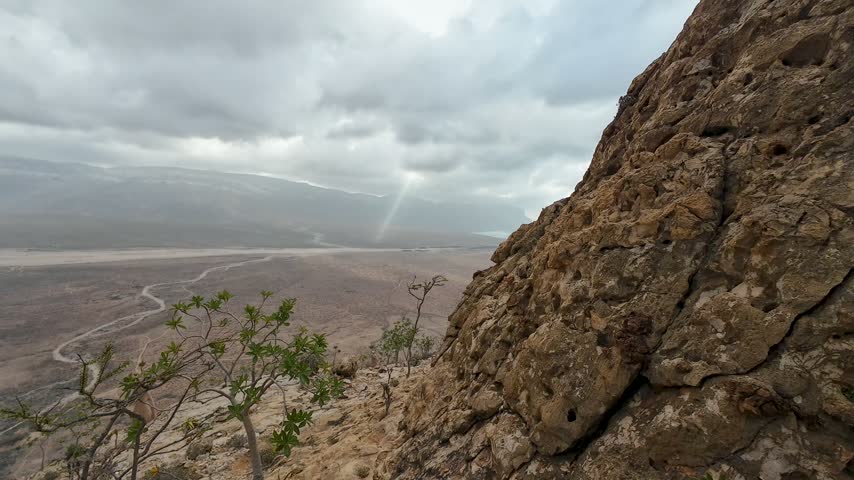 Dramatic storm clouds over a wide desert valley, viewed from a rocky mountain slope with sparse shrubs and soft sunlight breaking through. Socotra, Yemen.