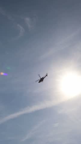 Vertical screen military helicopter flies near bright sun across a hazy blue sky during an aerial demonstration with spinning rotor blades and dramatic backlight overhead