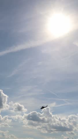 Vertical screen military helicopter flies beneath bright sun and dramatic clouds during aerial demonstration showing powerful rotorcraft motion across the open summer sky