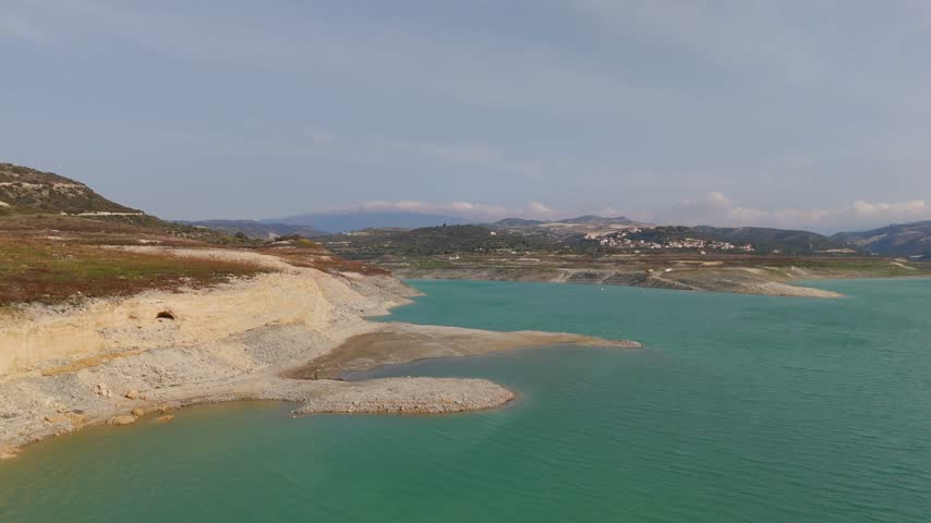 A low-altitude aerial tracking shot moves across the calm blue water of Kouris Dam toward a rocky, dry shoreline under bright daylight.