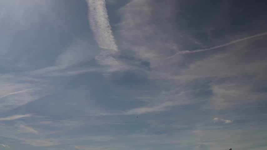 Distant fighter jet flies across a wide blue skies beneath layered white clouds and long vapor trails creating a dramatic aviation scene high above the ground in daylight