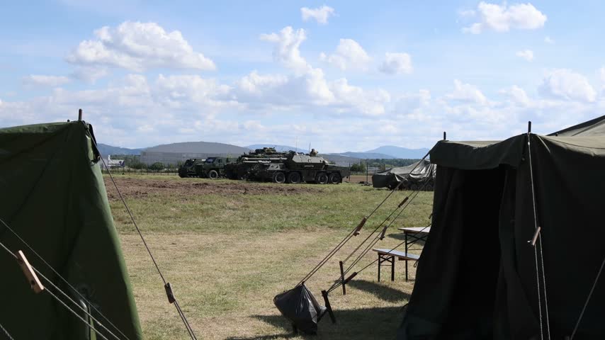 Military field camp with Pandur II 8x8 CZ armored personnel carrier and support vehicles parked between tents during an outdoor defense equipment display in daylight view