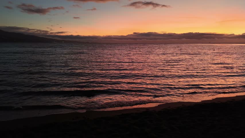 Aerial moving forward from Point Venus over rippling water, revealing the golden sunset horizon and dark coast of Tahiti island at twilight