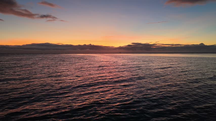 Aerial moving forward then backward from Point Venus over rippling water, revealing the golden sunset horizon and dark coast of Tahiti island at twilight