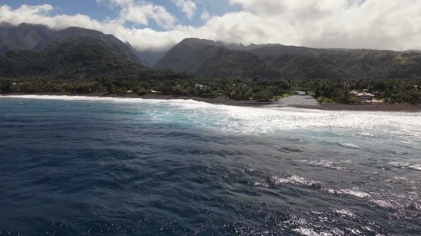 Aerial moving forward over white surf breaks, revealing the volcanic black sand beach, river mouth, and lush mountain valley of Taharuu Black Sand Beach in Tahiti
