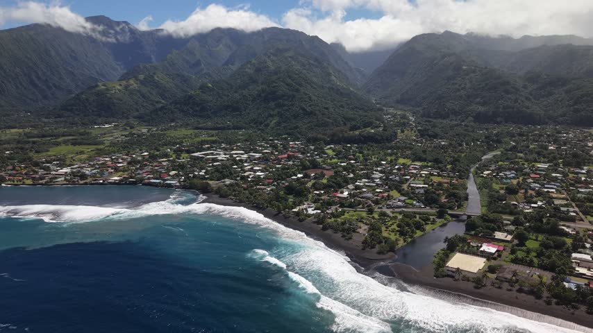 Wide stationary aerial showcasing the volcanic black sand beach, white surf breaks, river mouth, and lush mountain valley of Taharuu in Tahiti