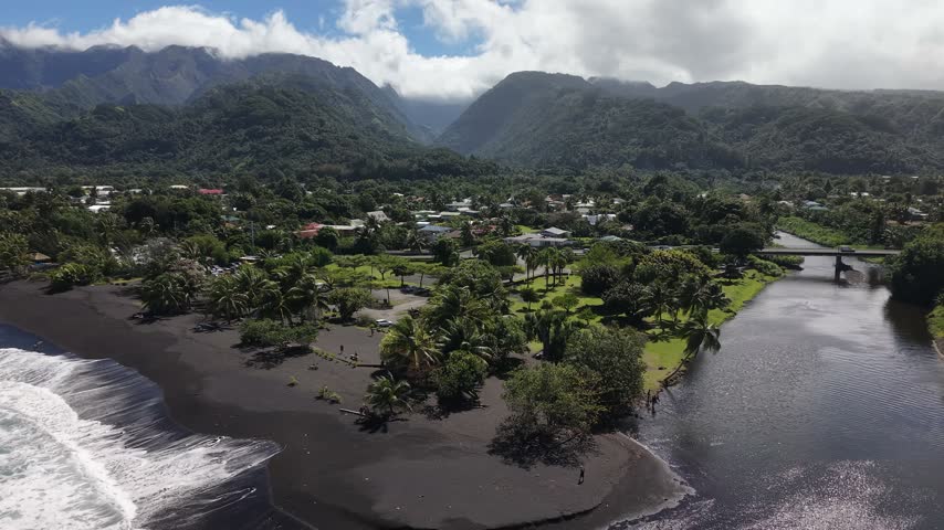 Wide high-angle aerial orbit shot showcasing a volcanic black sand beach, river mouth, coastal houses, and lush green mountains of Paea in Tahiti