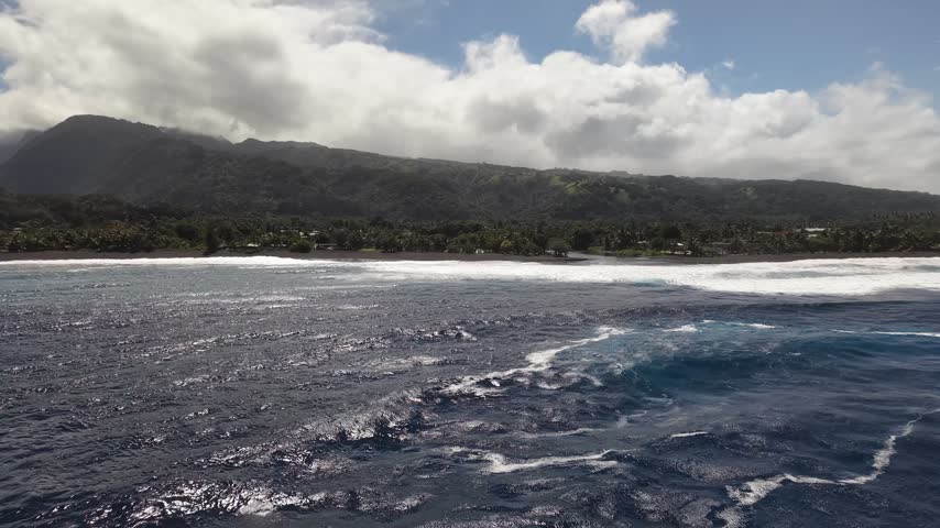 Aerial moving forward over white surf breaks, revealing the volcanic black sand beach, river mouth, and lush mountain valley of Paea in Tahiti