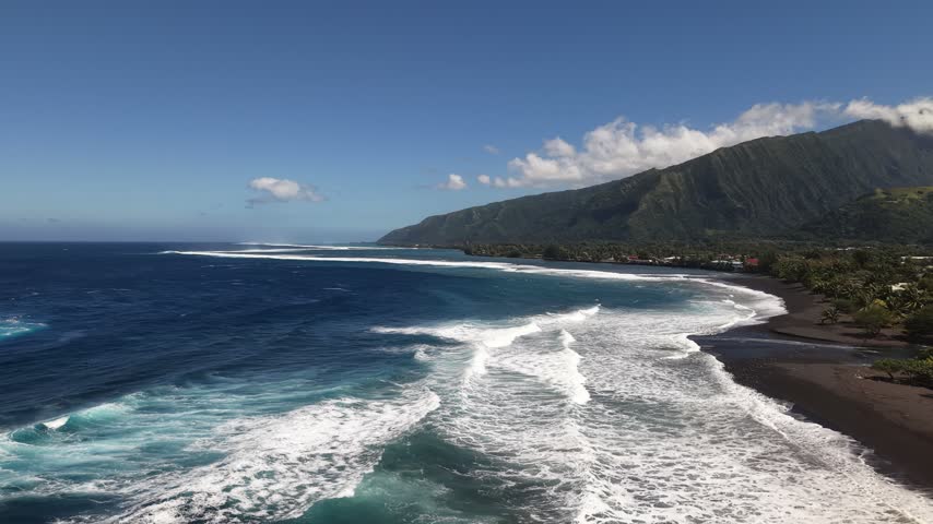 Aerial moving forward over white surf breaks, revealing the volcanic black sand beach, river mouth, and lush mountain valley of Paea in Tahiti