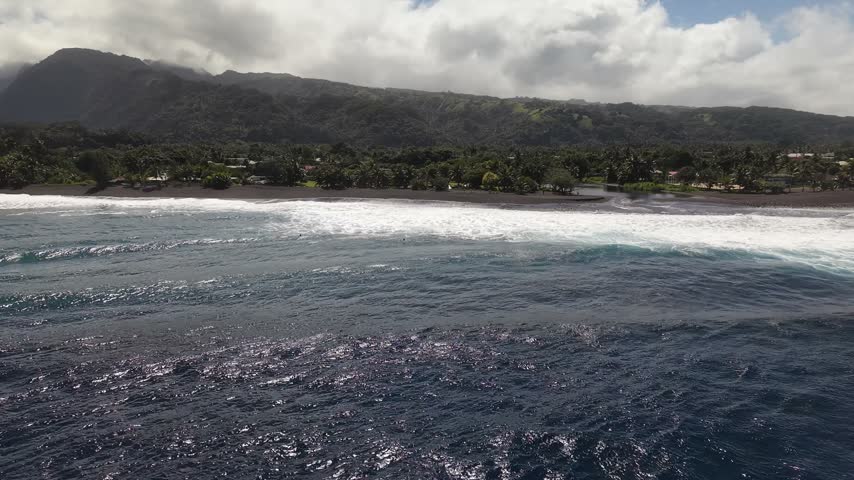 Aerial moving forward over white surf breaks, revealing the volcanic black sand beach, river mouth, and lush mountain valley of Paea in Tahiti