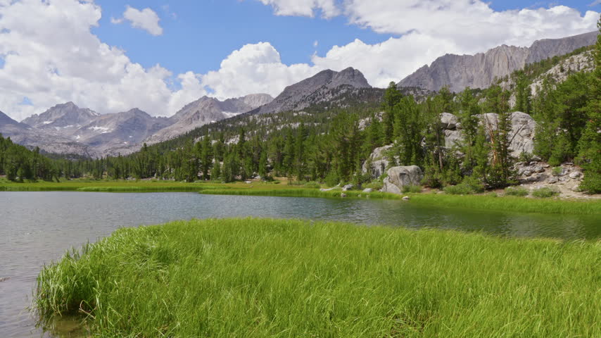A mountain lake bordered by meadow grasses and conifers beneath rugged granite peaks and passing clouds in the Sierra Nevada, California, United States.