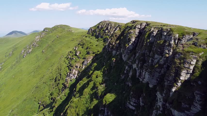 Aerial view of a group of hikers standing on top of a rocky mountain peak. Lush green slopes and a spectacular ridgeline under a clear blue sky