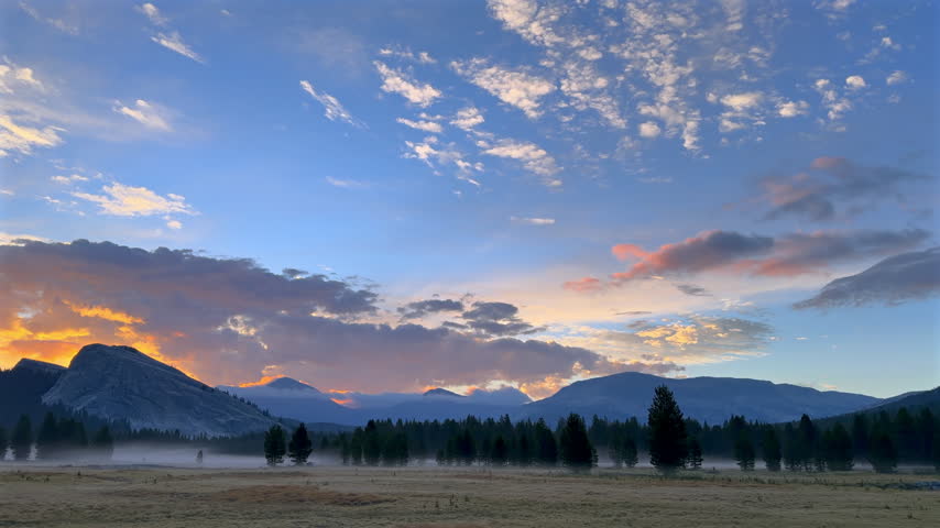 Warm sunrise clouds glow above misty Tuolumne Meadows, scattered conifers and distant peaks in Yosemite National Park, Sierra Nevada, California, United States.