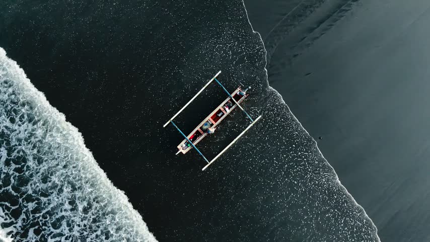 Top down drone view of traditional outrigger boat jukung surrounded by white foamy ocean waves on black sand beach. Dramatic action seascape finished vibrant color for cinematic film